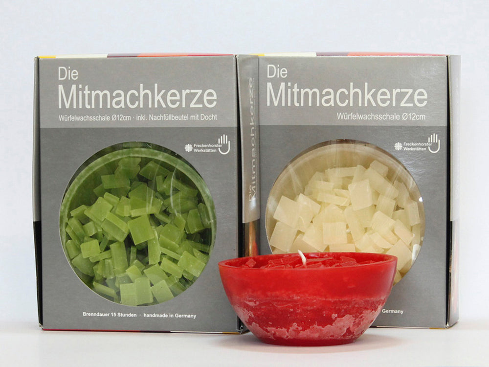 Rote Schalenkerze vor zwei Verpackungen „Die Mitmachkerze“ mit grünem und weißem Wachsgranulat, freigestellt auf Weiß.