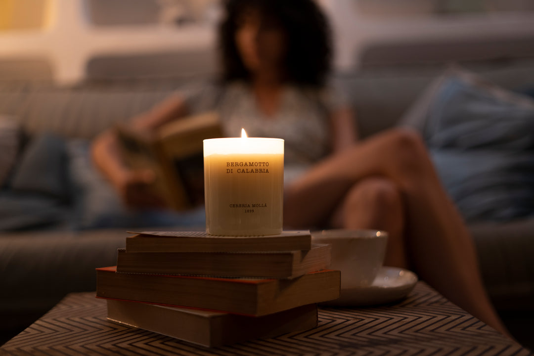 Brennende Duftkerze „Bergamotto di Calabria“ auf einem Bücherstapel, im Hintergrund eine entspannte Frau auf dem Sofa mit Buch – stimmungsvolles Abendambiente