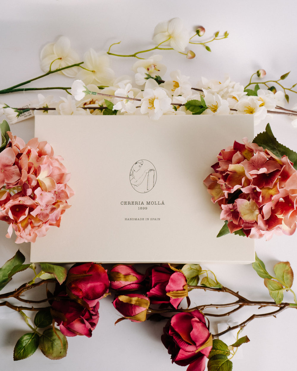Elegante Geschenkbox von Cereria Mollá mit feinem Logoaufdruck, umgeben von zarten Orchideen, Hortensien und Rosen. Die florale Komposition vermittelt Stil, Hochwertigkeit und liebevolle Handarbeit – ideal als Präsent oder Dekoration