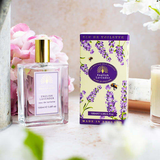 „Eau de Toilette ‘English Lavender’ in Glasflasche mit goldener Kappe, daneben passende Kartonverpackung mit Lavendelmotiv, im Hintergrund Dekoblumen.“