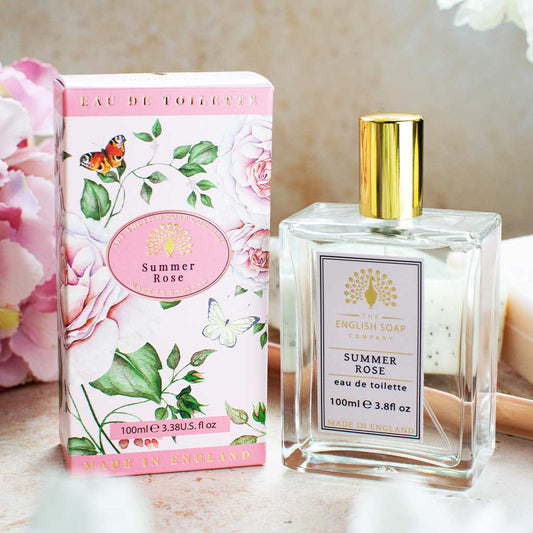 „Flasche und rosafarbene Verpackung der Eau de Toilette ‘Summer Rose’ auf hellem Untergrund mit Blumen im Hintergrund arrangiert.“