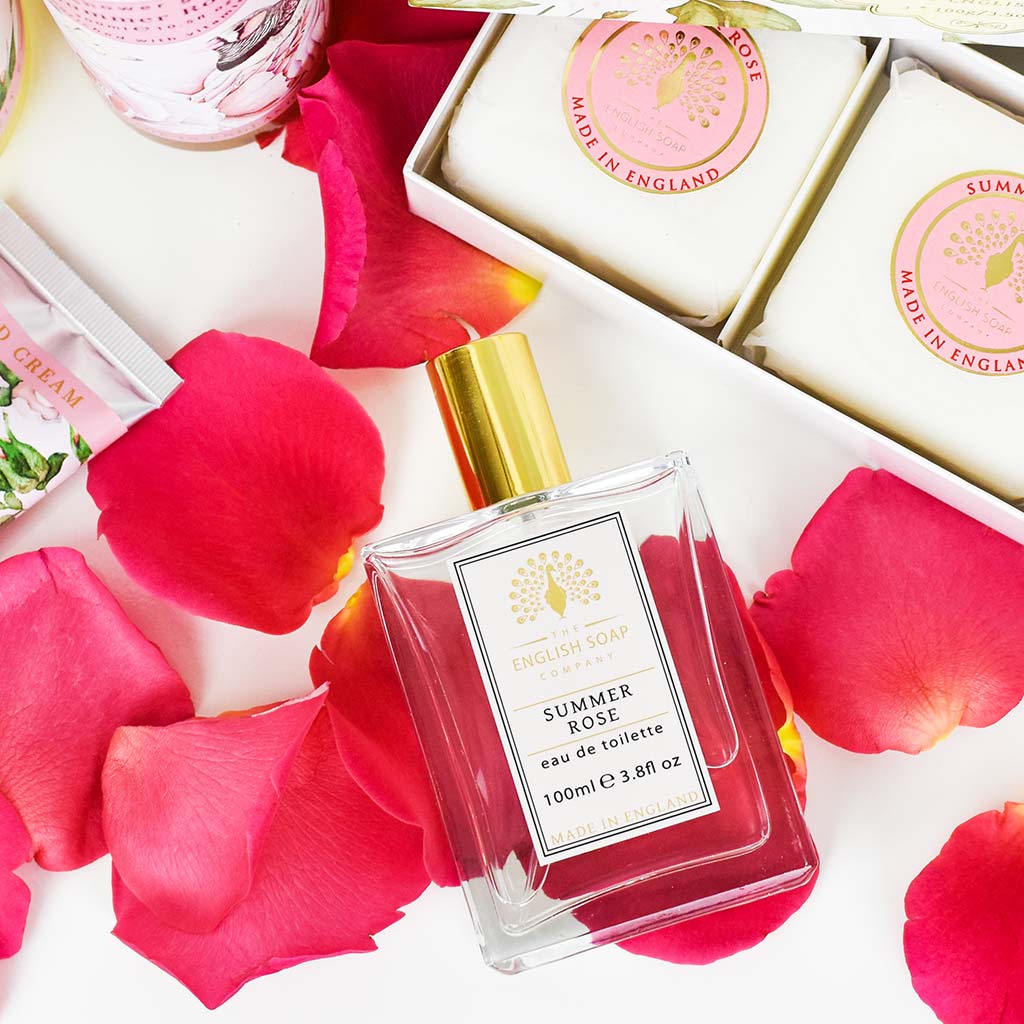 „Flasche der Eau de Toilette ‘Summer Rose’ mit goldener Verschlusskappe auf weißen Hintergrund und roten Rosenblättern, daneben Seifenstücke in Geschenkverpackung.“