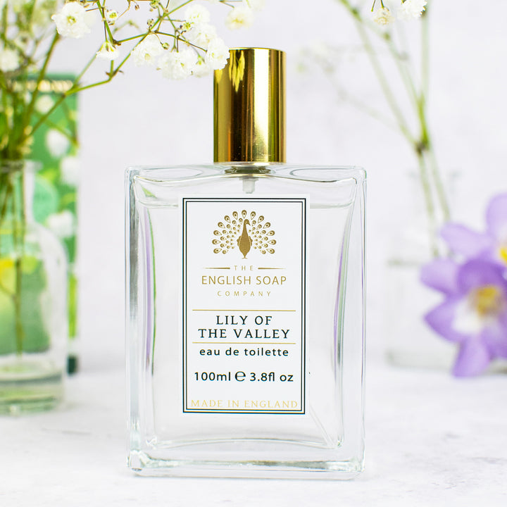 „Flakon der Eau de Toilette ‘Lily of the Valley’ mit goldener Verschlusskappe, auf weißem Untergrund vor Glasvasen mit Blumen arrangiert.“