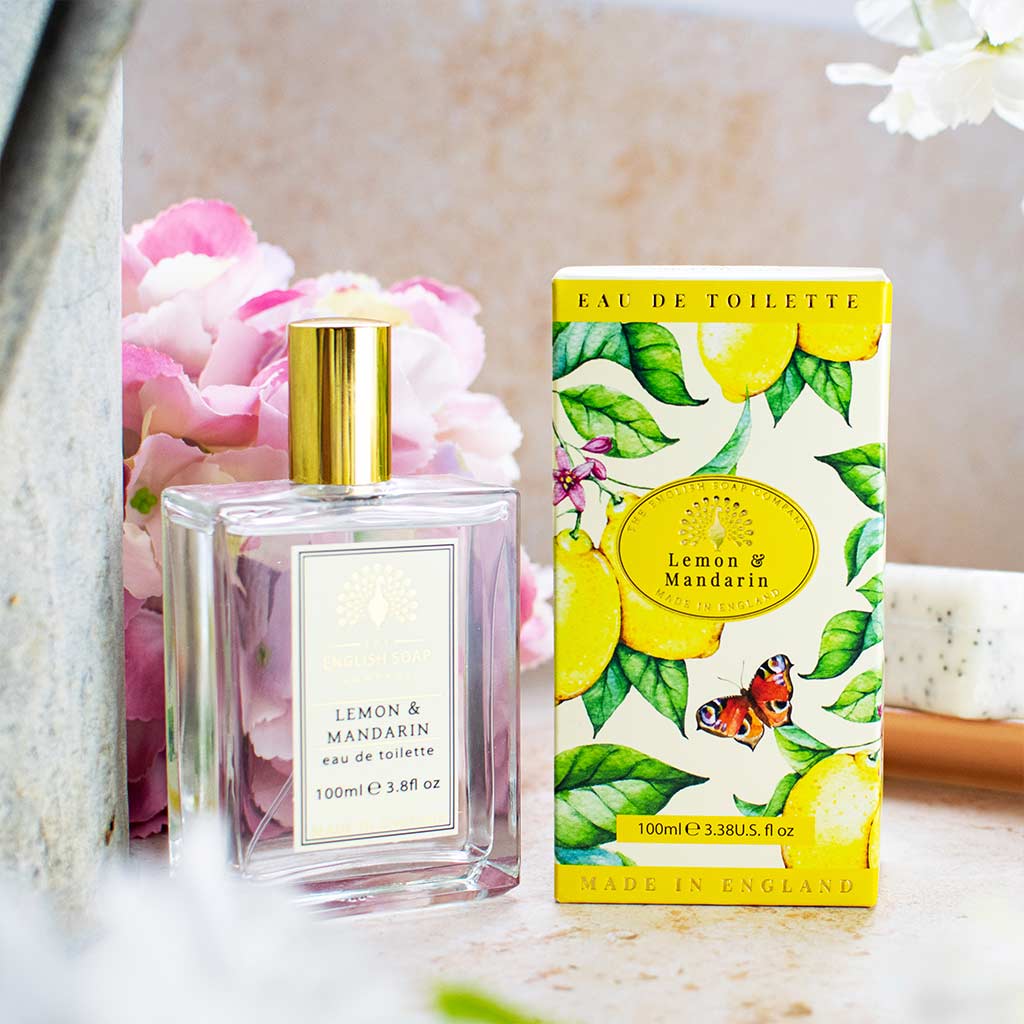 „Eau de Toilette ‘Lemon & Mandarin’ mit goldener Verschlusskappe und floraler Verpackung mit gelbem Etikett, arrangiert vor rosa Blüten.“