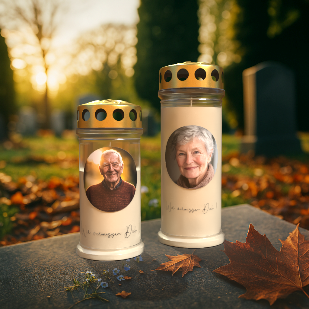 Zwei Gedenkkerzen mit goldfarbenen Deckeln stehen auf einem Grabstein auf einem Friedhof bei Sonnenuntergang. Auf der linken Kerze ist das Porträt eines älteren Mannes abgebildet, auf der rechten das einer älteren Frau. Beide Kerzen tragen die Inschrift „Wir vermissen Dich“. Im Vordergrund liegen herbstliche Blätter und kleine blaue Blumen, während im Hintergrund Grabsteine und Bäume im warmen Licht der untergehenden Sonne zu sehen sind.