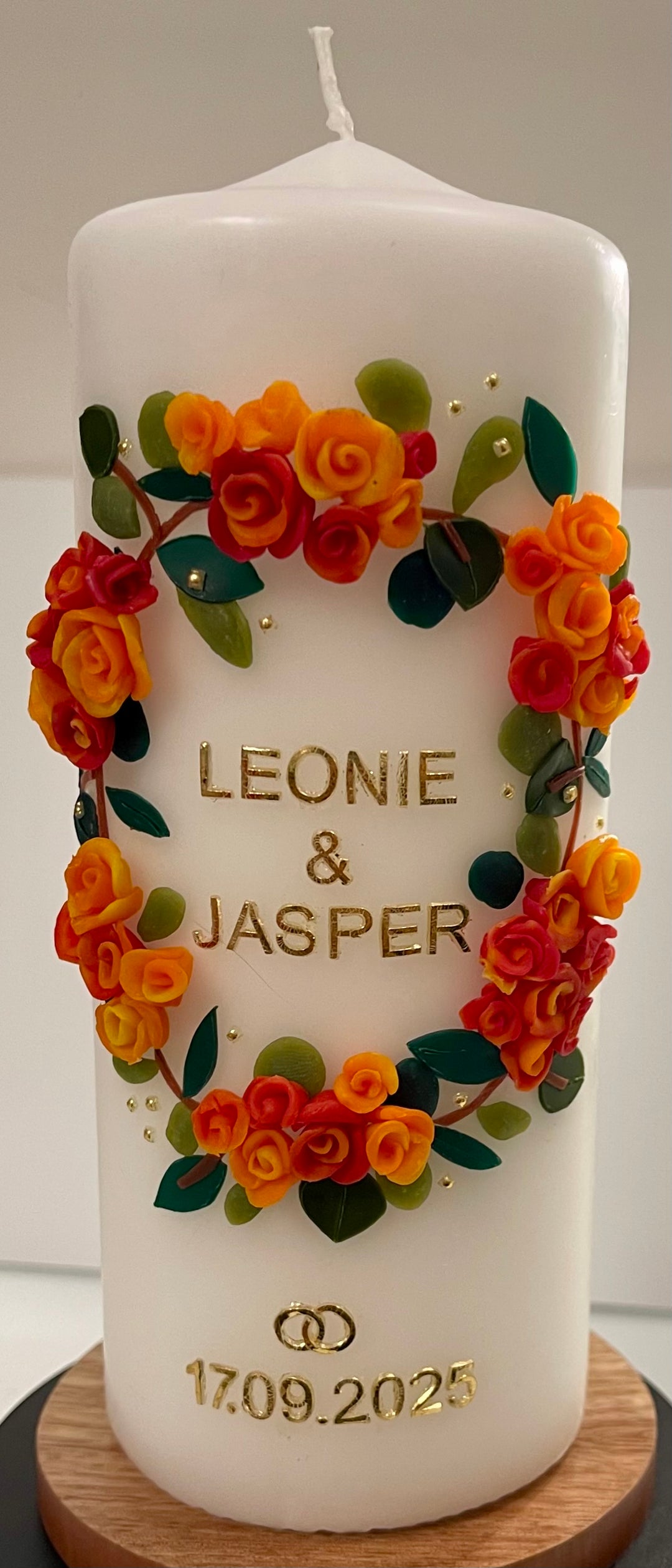 Weiße Hochzeitskerze mit handgefertigtem Wachsblumenkranz in Gelb, Orange und Rot, goldener Aufschrift „Leonie & Jasper“, goldenen Ringen und Hochzeitsdatum 17.09.2025, auf Holzsockel präsentiert.
