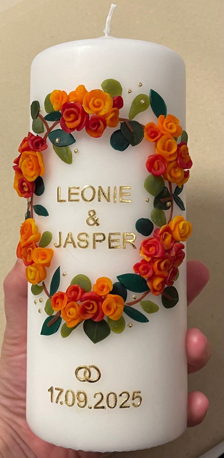 Weiße Hochzeitskerze mit handgefertigtem Wachsblumenkranz in Gelb, Orange und Rot, goldener Aufschrift ‚Leonie & Jasper‘, goldenen Ringen und Hochzeitsdatum 17.09.2025