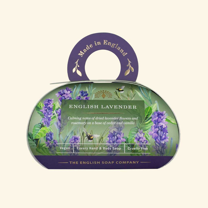 „Luxus-Hand- und Körperseife der Marke The English Soap Company in ovaler Verpackung mit Tragegriff, Motiv: Lavendelblüten, Bienen und Kräuter auf hellem Hintergrund, Aufschrift: ‚English Lavender – Calming notes of dried lavender flowers and rosemary on a base of cedar wood and vanilla‘.“