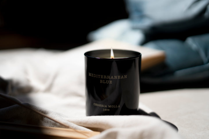 Schwarze Duftkerze Mediterranean Blue brennt in sanftem Licht auf einer weichen Stoffunterlage. Im Hintergrund liegen gefaltete blaue Kissen – die Szene wirkt ruhig, cozy und maritim inspiriert
