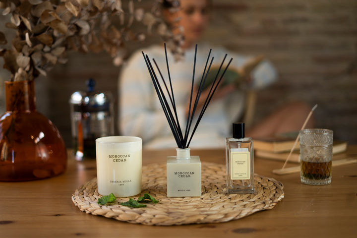Moroccan Cedar Duftkollektion von Cereria Mollá auf einem Holztisch: Duftkerze, Raumduft mit Stäbchen und Parfumflakon elegant arrangiert auf einem geflochtenen Untersetzer. Im Hintergrund eine entspannt sitzende Frau mit warmer Teetasse – stilvolle Wohlfühlatmosphäre mit orientalischem Touch