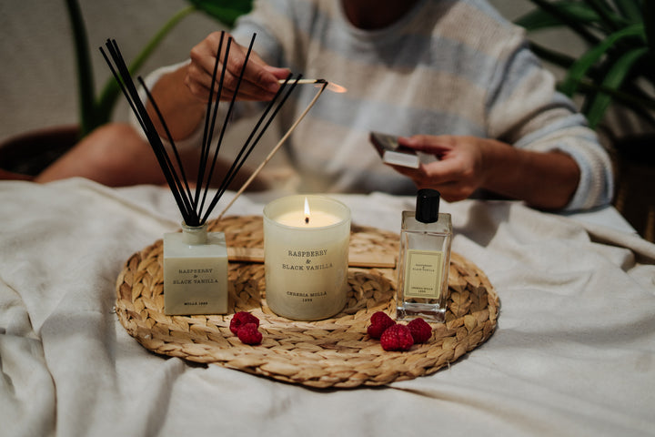 Duftkerze Raspberry & Black Vanilla von Cereria Mollá auf einem Tablett mit Raumduft, Diffusor-Stäbchen, Himbeeren und gemütlichem Hintergrund – eine Hand zündet die Kerze an.