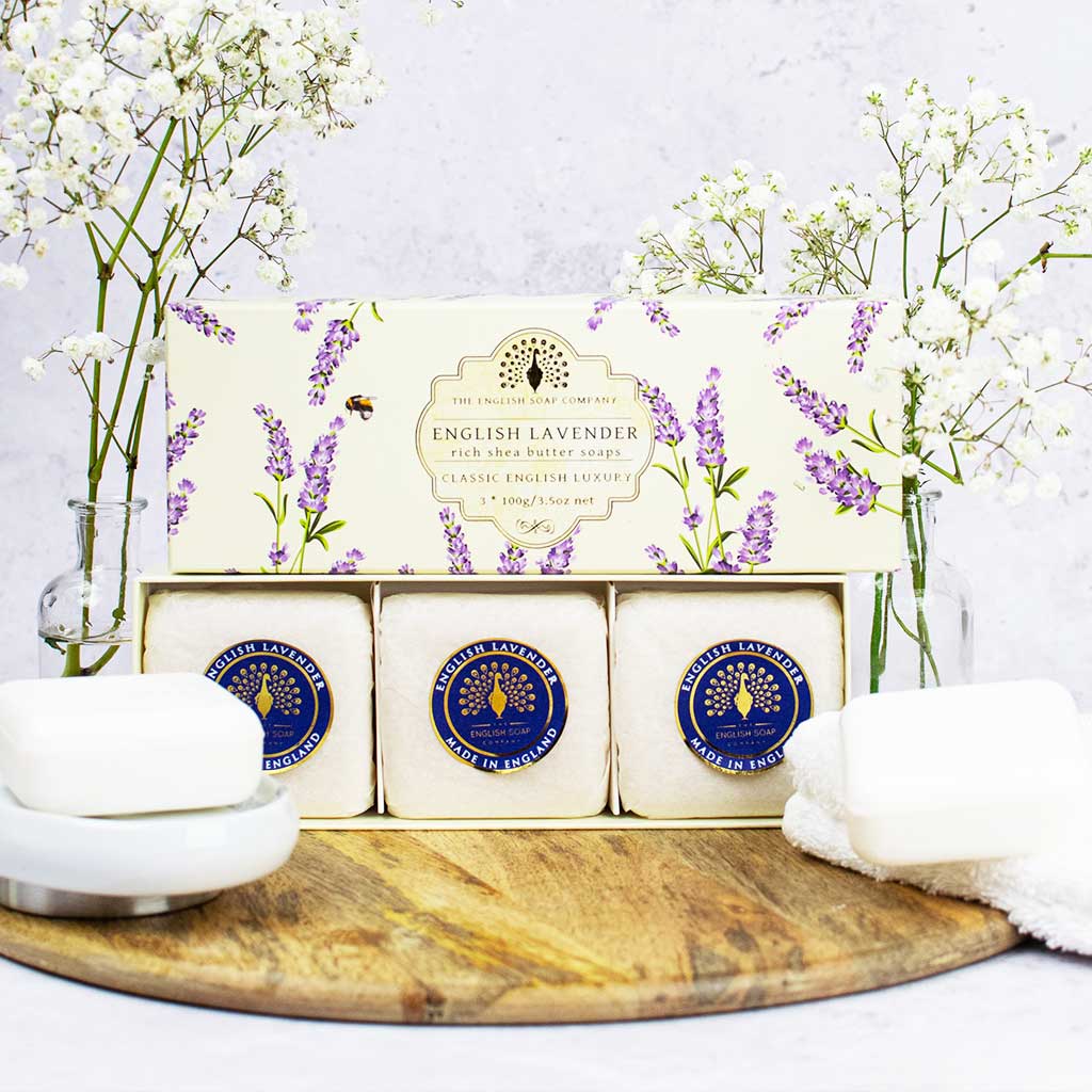 „Geschenkbox mit drei runden Seifen der Sorte ‘English Lavender’ auf Holzunterlage, dekoriert mit Schleierkraut und Handtuch.“