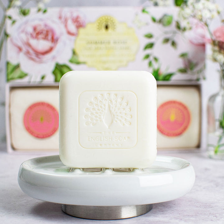 „Ein Stück der ‘Summer Rose’-Seife mit Prägung auf einer weißen Seifenschale, im Hintergrund die geöffnete Geschenkbox mit weiteren Seifenstücken und floraler Verpackung.“