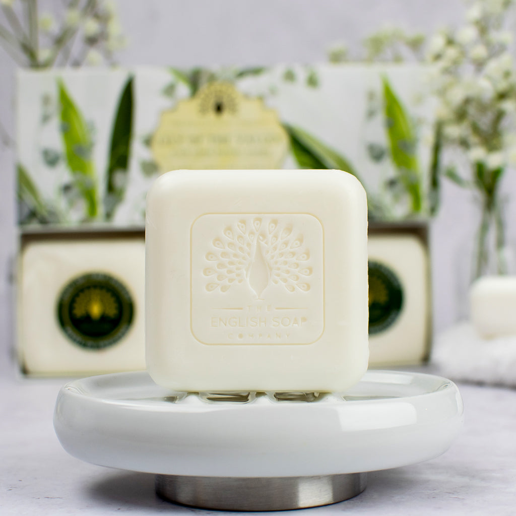 „Einzelnes Seifenstück der Sorte ‘Lily of the Valley’ mit Prägung auf einer weißen Seifenschale, im Hintergrund Geschenkbox und dekorative Blumen.“