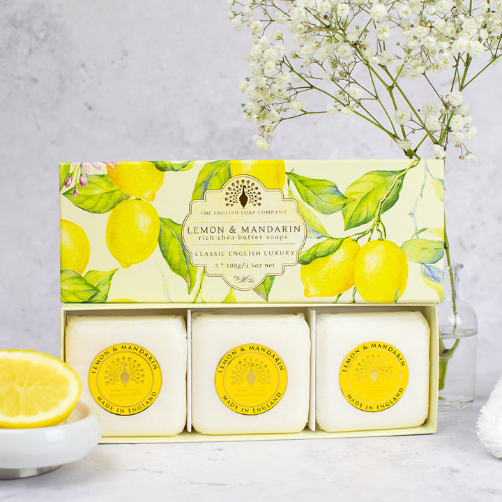 „Geschenkbox mit drei runden Seifen der Sorte ‘Lemon & Mandarin’ vor einer Verpackung mit gelben Zitronenmotiven, dekoriert mit Blütenzweig und Zitronenscheibe.“