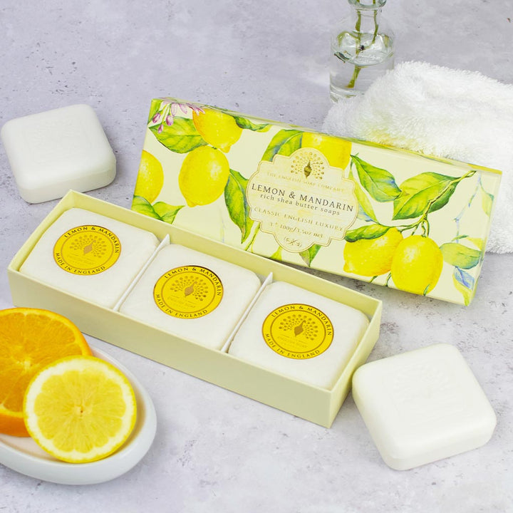 „Geschenkbox mit drei runden Seifen der Sorte ‘Lemon & Mandarin’ neben zwei ausgepackten Seifenstücken, Zitrusscheiben und einem Handtuch auf heller Oberfläche.“