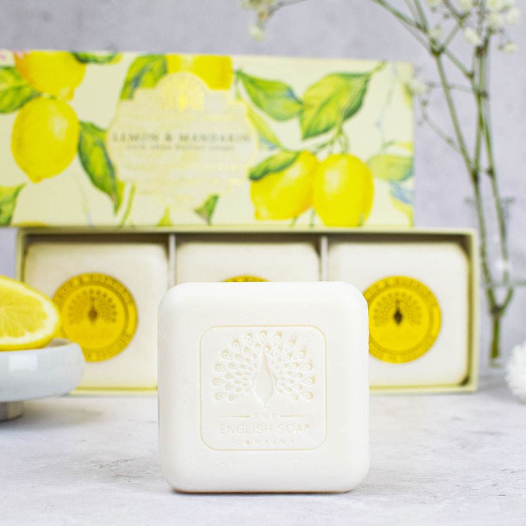 „Nahaufnahme eines ausgepackten Seifenstücks mit Prägung vor einer Geschenkbox mit drei runden Seifen der Sorte ‘Lemon & Mandarin’ und einer aufgeschnittenen Zitrone im Hintergrund.“