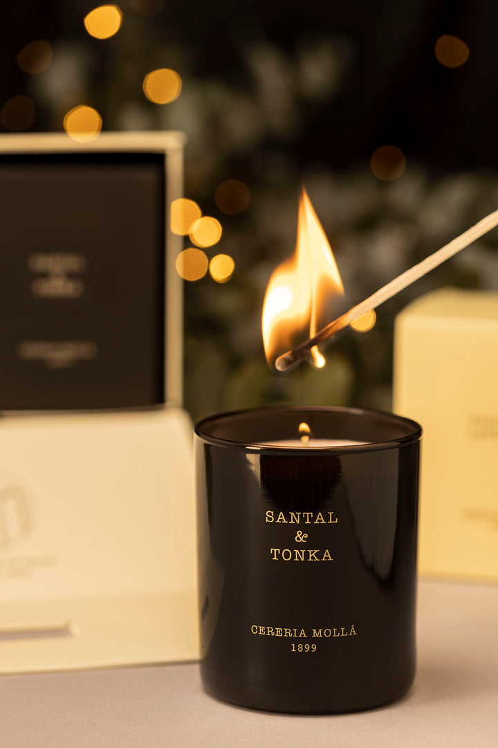 Eine schwarze Duftkerze mit goldener Schrift (SANTAL & TONKA – CERERIA MOLLA 1899) wird mit einem brennenden Streichholz angezündet. Im Hintergrund stimmungsvolle Lichtreflexe und elegante Verpackungen – festliche, luxuriöse Atmosphäre