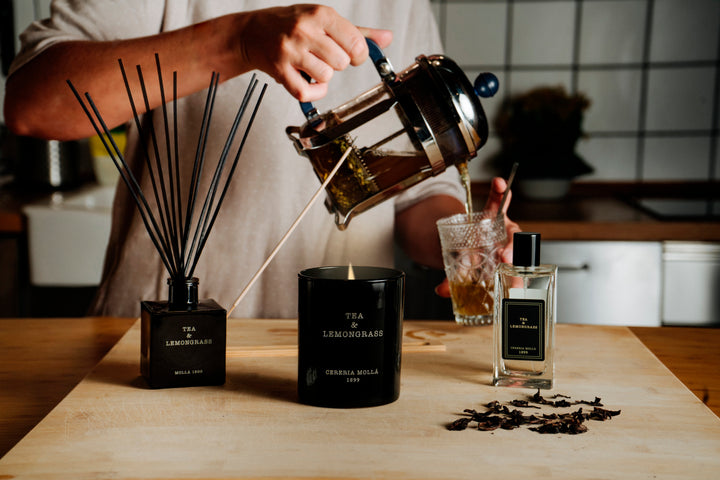 Stimmungsvolles Küchen- oder Wohnambiente mit der Duftkerze Tea & Lemongrass von Cereria Mollá, einem passenden Raumduft und einem Teeglas; im Vordergrund getrocknete Teeblätter und eine Hand, die Tee eingießt
