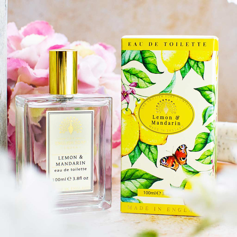 Eau de Toilette Lemon & Mandarin mit goldener Verschlusskappe, transparentem Flakon und floraler Verpackung mit Zitrusmotiv, arrangiert vor rosa Blüten.
