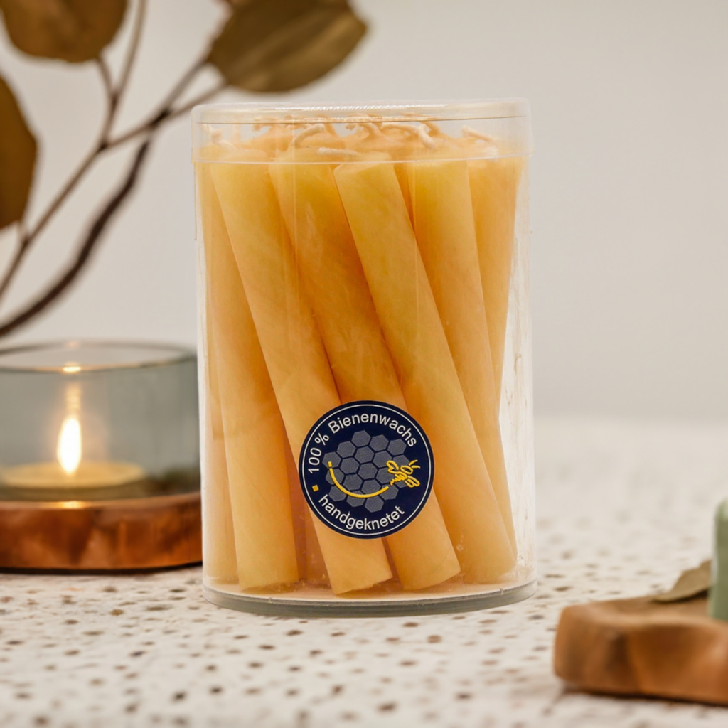 Glas mit mehreren dünnen Christbaumkerzen aus Bienenwachs, dekorativ platziert auf einem hellen Untergrund mit Kerze im Hintergrund