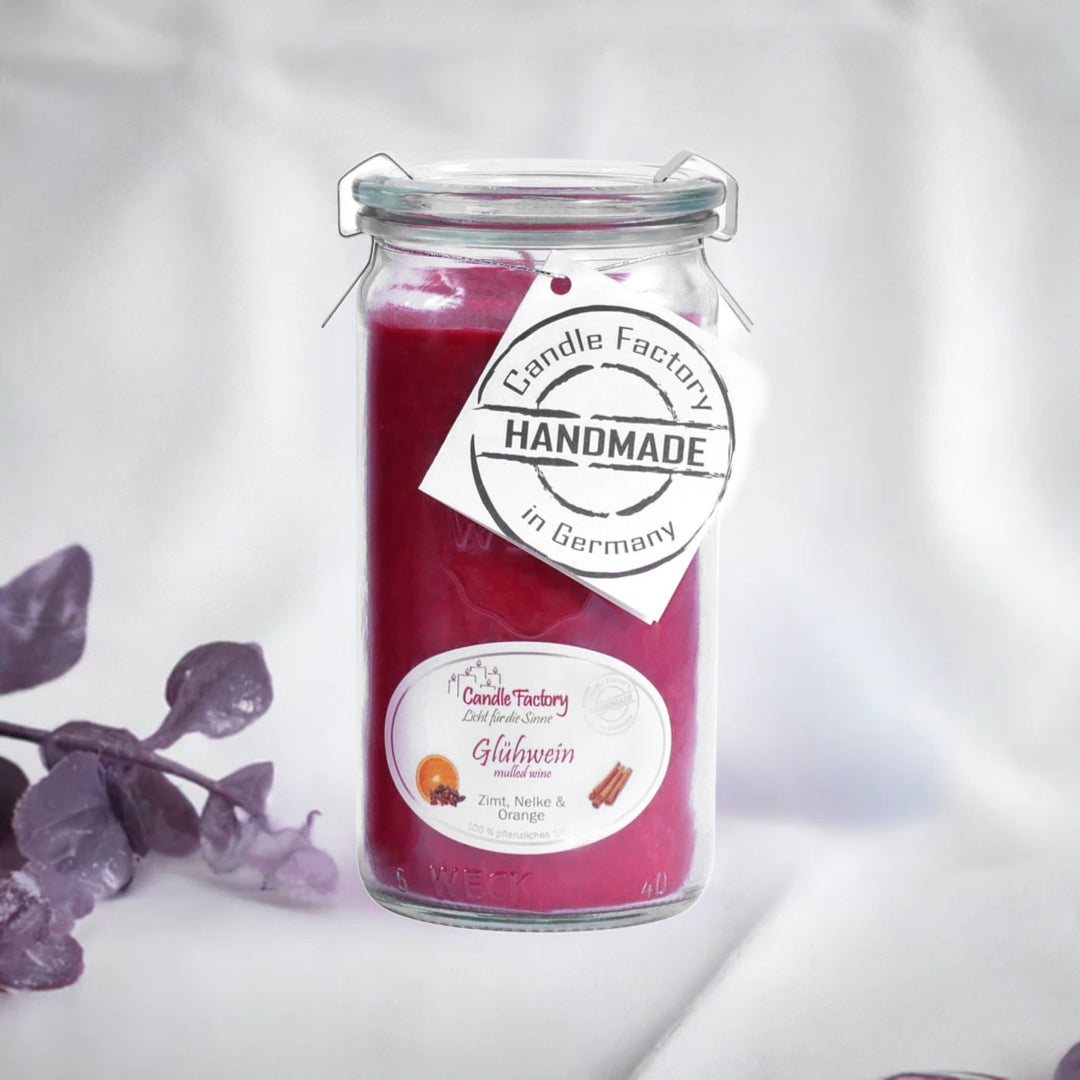 Rote Duftkerze im Glas mit Etiketten „Glühwein“ und „Handmade in Germany“, daneben violette Dekozweige auf hellem Hintergrund.