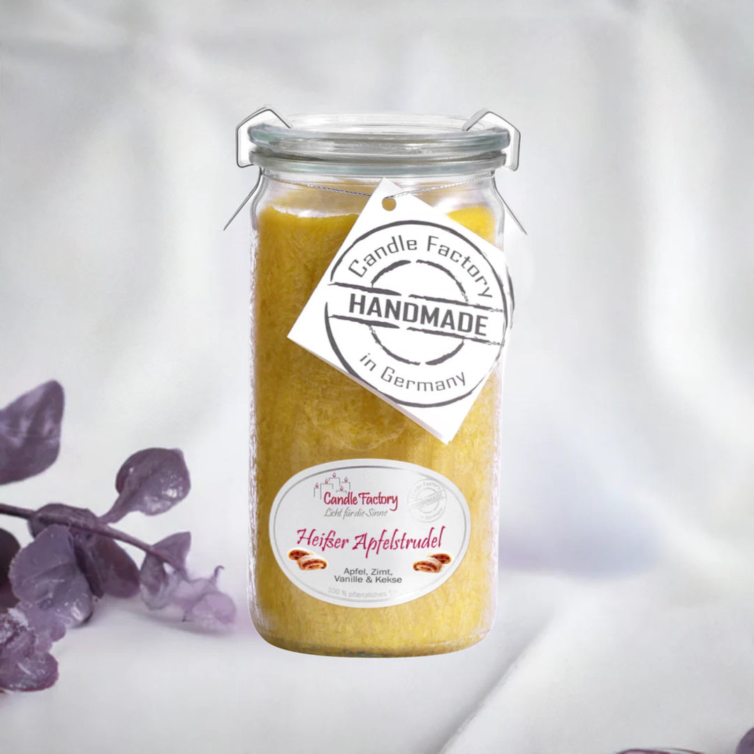 Gelbe Duftkerze im Glas mit Etiketten „Heißer Apfelstrudel“ und „Handmade in Germany“, daneben violette Dekozweige.