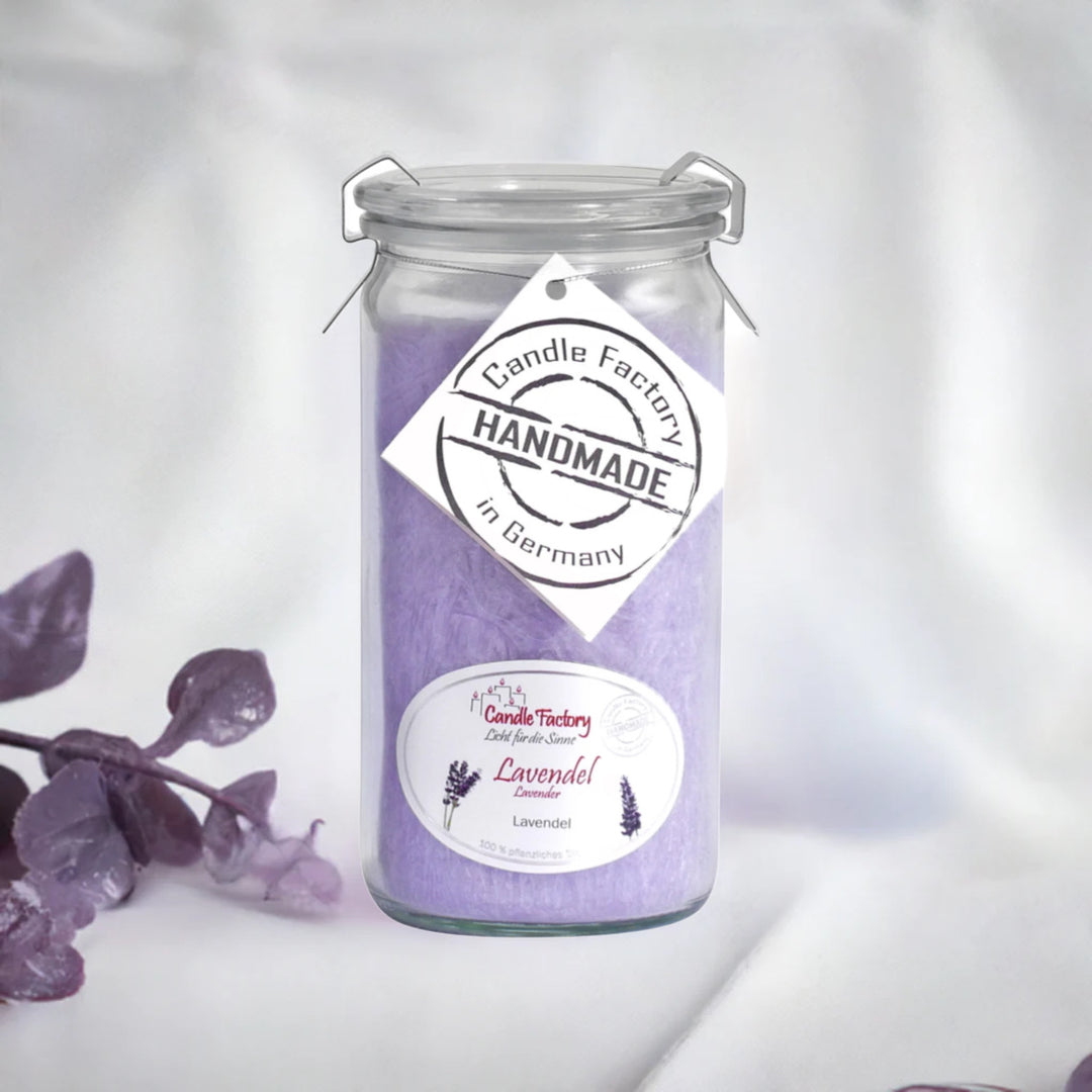 Lavendelfarbene Duftkerze im Glas mit „Handmade in Germany“-Etikett, dekorativ platziert neben lila Blättern auf hellem Stoff.