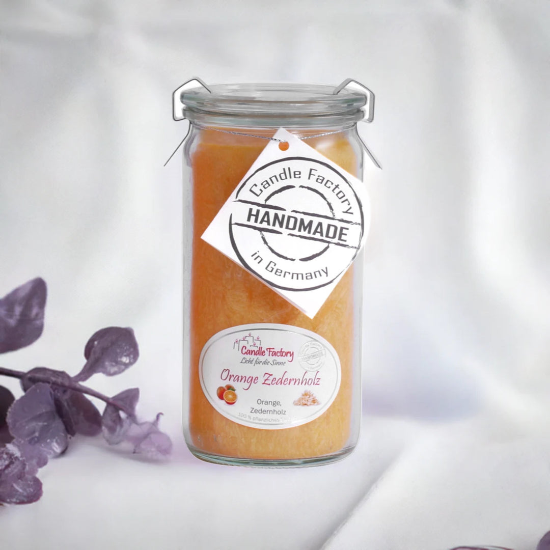 Orange Duftkerze im Glas mit Etiketten „Orange Zedernholz“ und „Handmade in Germany“, daneben violette Dekozweige.