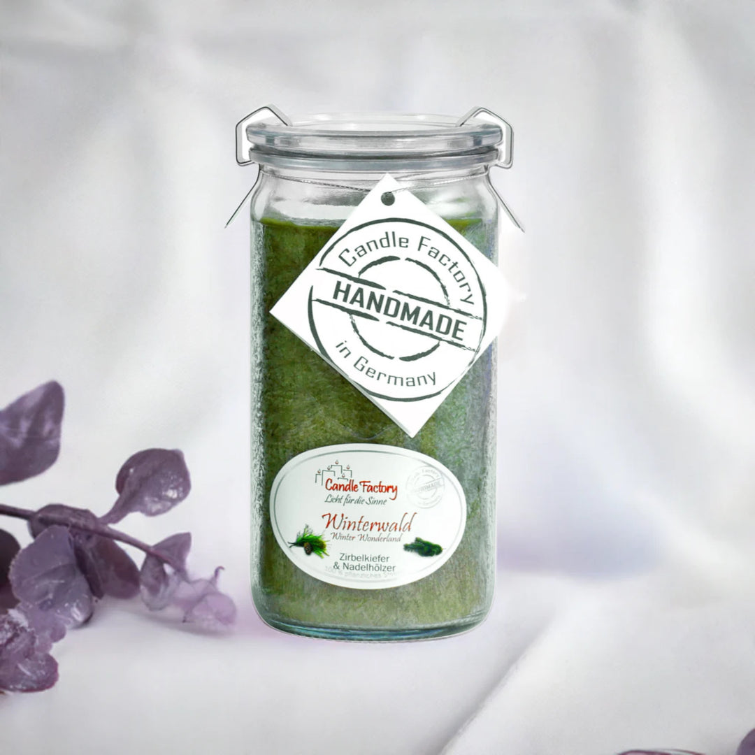 Grüne Duftkerze im Glas mit Etiketten „Winterwald“ und „Handmade in Germany“, daneben violette Dekozweige auf hellem Hintergrund.