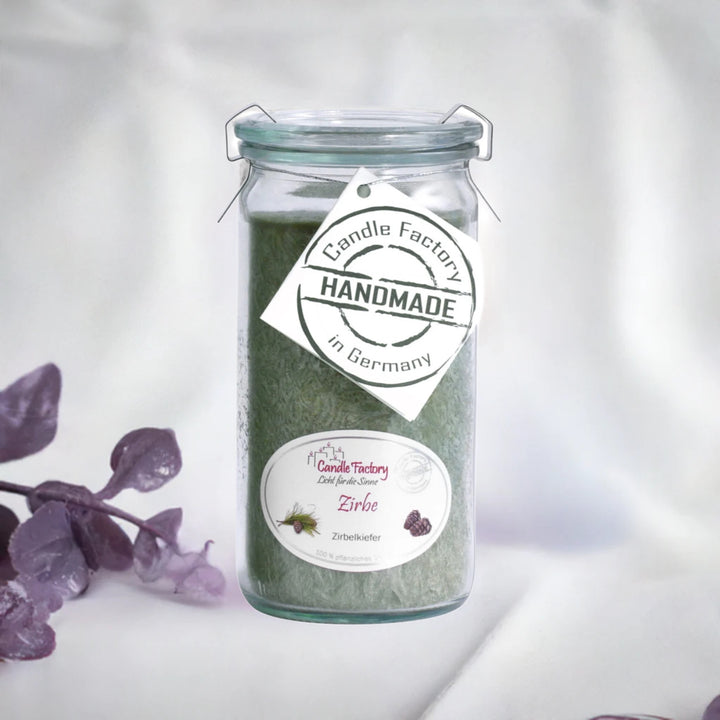Grüne Duftkerze im Glas mit Etiketten „Zirbe“ und „Handmade in Germany“, daneben violette Dekozweige auf hellem Hintergrund.