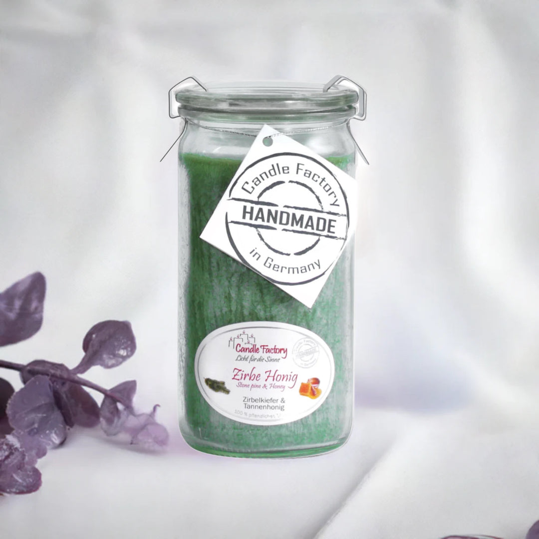 Grüne Duftkerze im Glas mit Etiketten „Zirbe-Honig“ und „Handmade in Germany“, daneben violette Dekozweige auf hellem Hintergrund.