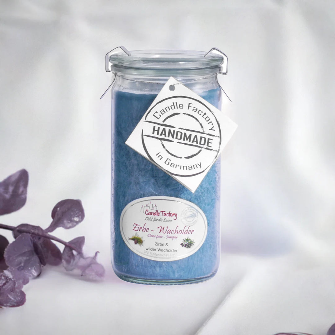 Blaue Duftkerze im Glas mit Etikett „Zirbe-Wacholder“ und „Handmade“, daneben violette Dekozweige auf hellem Hintergrund.