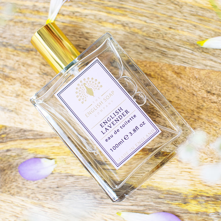 Flasche der Eau de Toilette „English Lavender“ mit goldener Verschlusskappe, liegend auf einer Holzoberfläche mit verstreuten Blütenblättern.