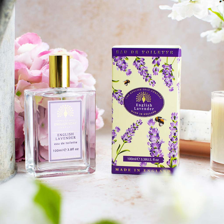 Eau de Toilette „English Lavender“ in Glasflasche mit goldener Kappe, daneben passende Kartonverpackung mit Lavendelmotiv, im Hintergrund Dekoblumen.