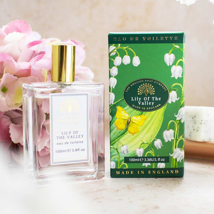 Flakon und grüne Verpackung des Eau de Toilette „Lily of the Valley“ neben rosafarbenen Blumen auf weißem Untergrund arrangiert.
