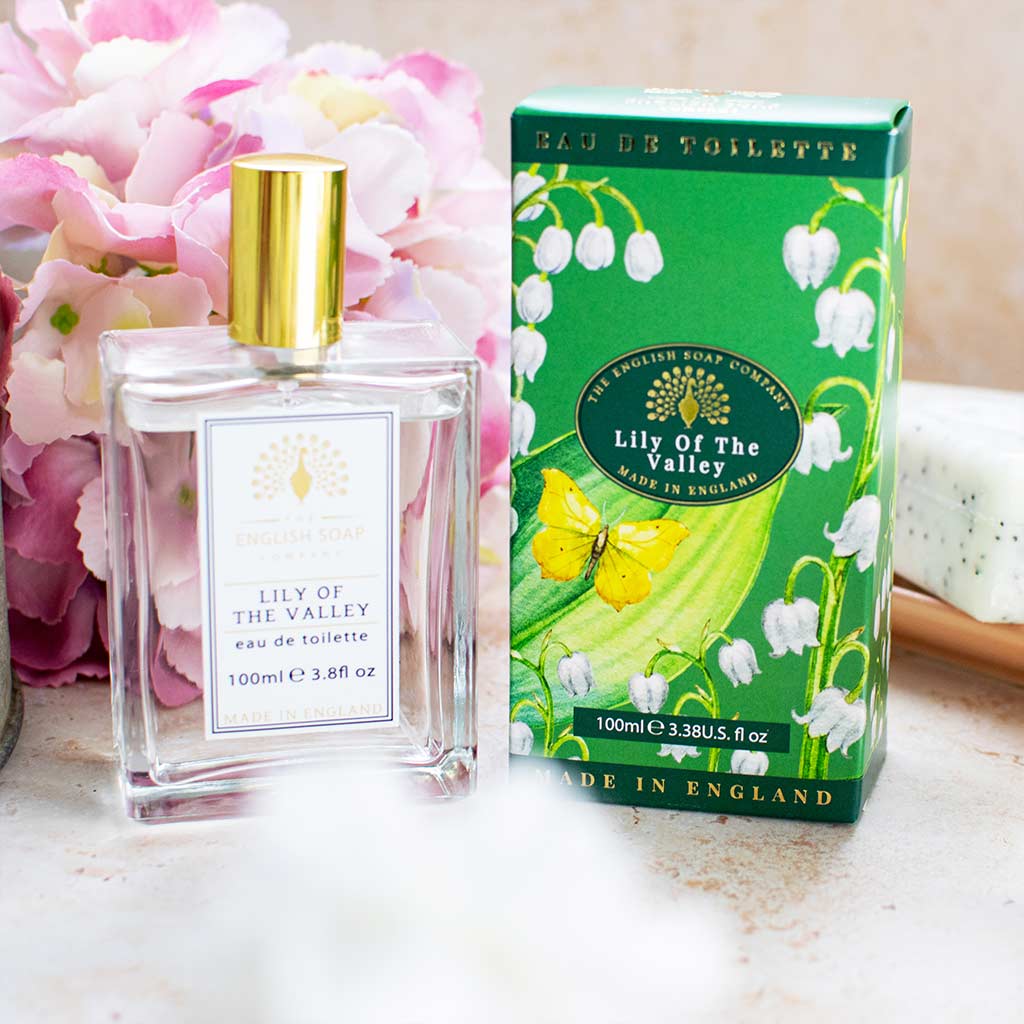 Eau de Toilette „Lily of the Valley“ mit goldener Verschlusskappe und grün-weißer Verpackung mit Maiglöckchenmotiv, arrangiert vor rosa Blüten.