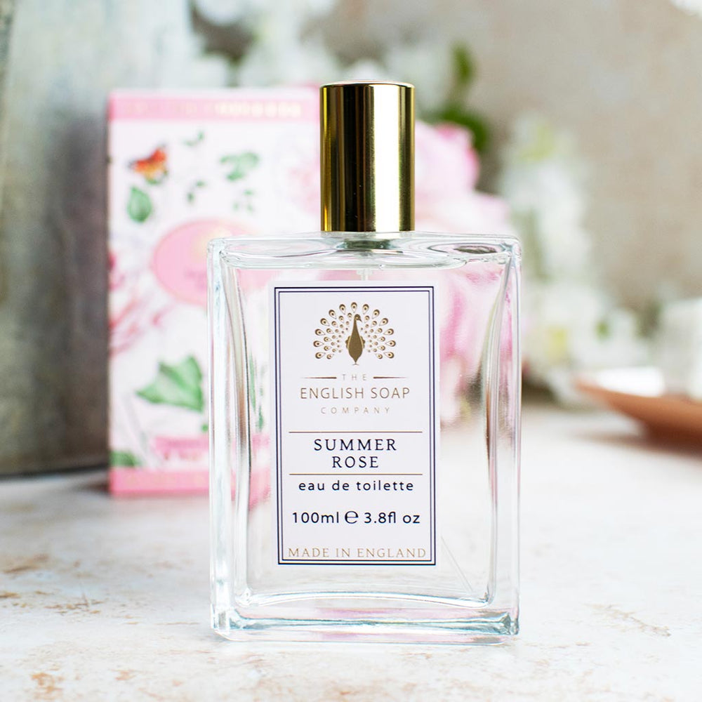 Eau de Toilette „Summer Rose“ im Glasflakon mit goldener Kappe, ohne Verpackung, auf hellem Untergrund mit zarten Blüten im Hintergrund arrangiert.