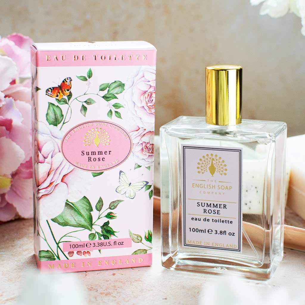 Flasche und rosafarbene Verpackung der Eau de Toilette „Summer Rose“ auf hellem Untergrund mit Blumen im Hintergrund arrangiert.