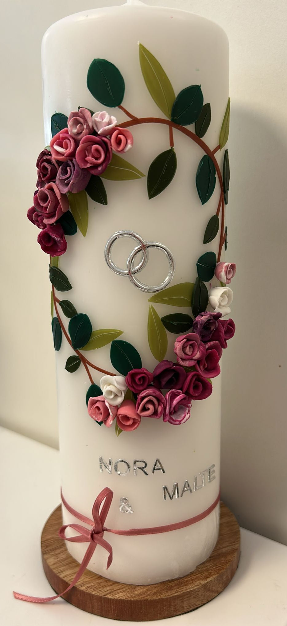 Weiße Hochzeitskerze mit handgefertigtem Wachsblumenkranz in Rosa-, Pink- und Lilatönen mit grünen Blättern, silbernen Ringen, silberner Aufschrift „Nora & Malte“ und rosa Schleifenband am unteren Rand.