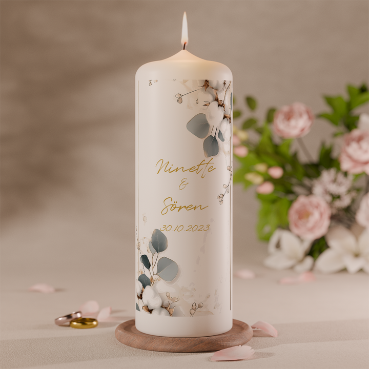 Hochzeitskerze mit Eukalyptusmotiv in Grün und Gold personalisiert mit Namen und Datum vor Blumen
