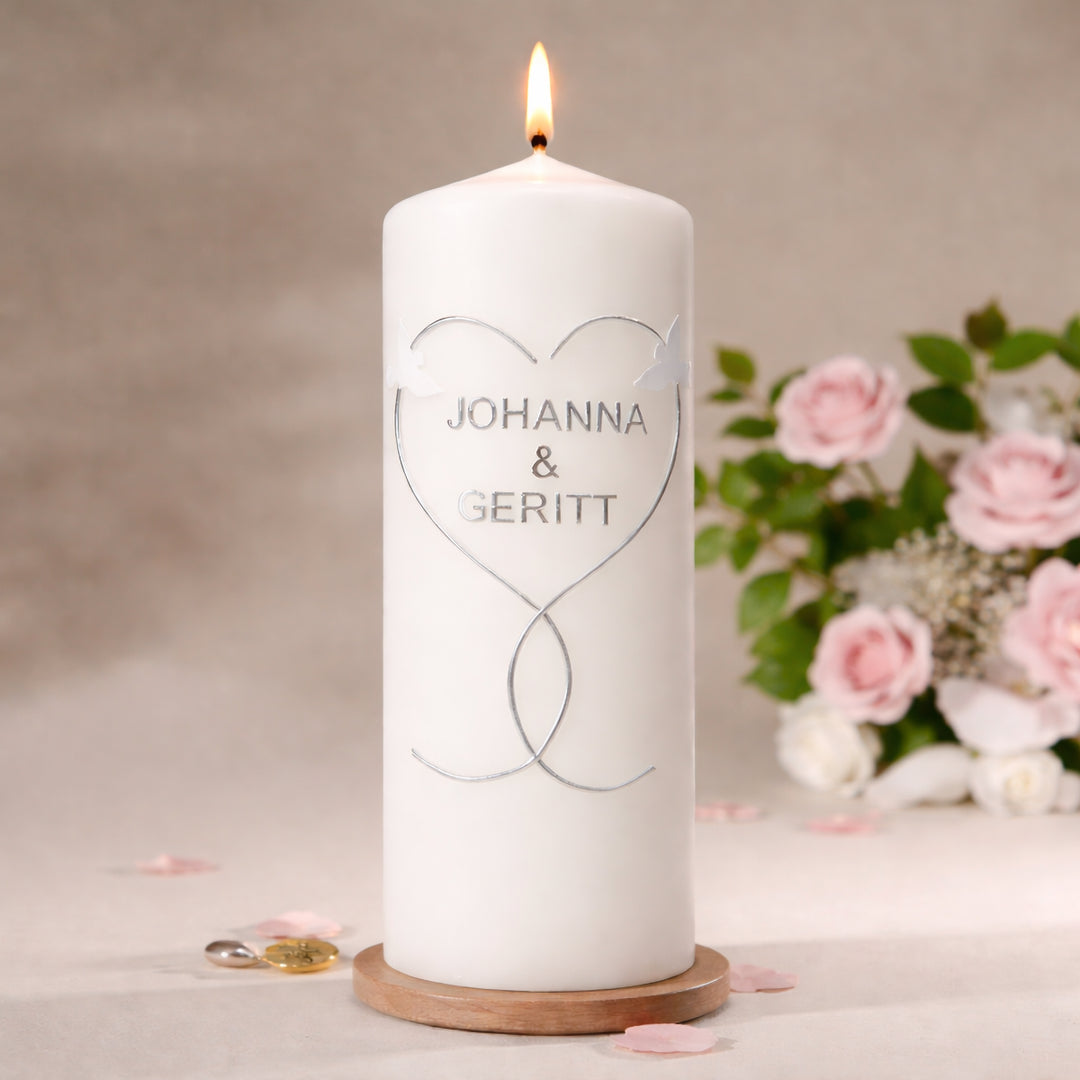 Handverzierte Hochzeitskerze in weiß mit zwei weißen Tauben und silbernem Herzmotiv auf Stumpenkerze, dekoriert auf Holzuntersetzer mit Blumen im Hintergrund
