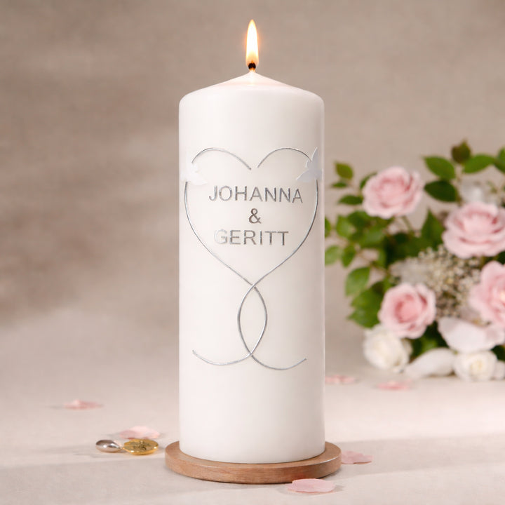 Handverzierte Hochzeitskerze in weiß mit zwei weißen Tauben und silbernem Herzmotiv auf Stumpenkerze, dekoriert auf Holzuntersetzer mit Blumen im Hintergrund

