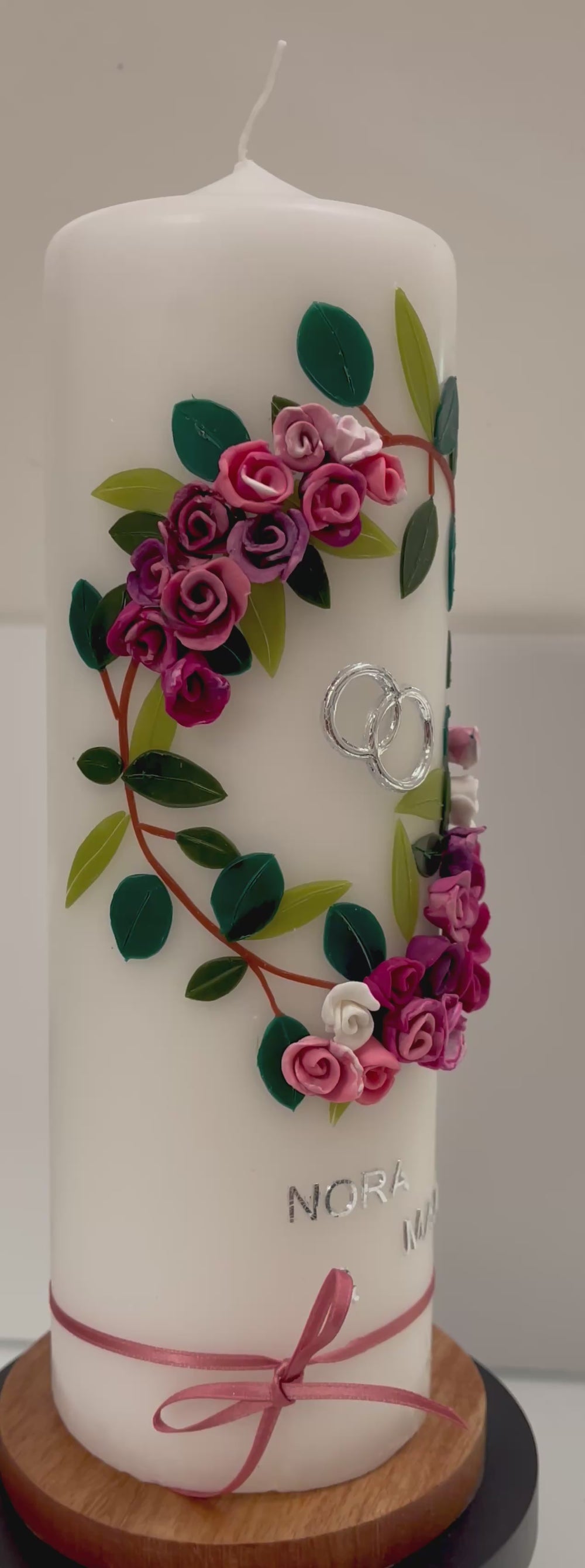Weiße Hochzeitskerze mit handgefertigtem Wachsblumenkranz in Rosa-, Pink- und Lilatönen mit grünen Blättern, silbernen Ringen, silberner Aufschrift „Nora & Malte“ und rosa Schleifenband am unteren Rand.