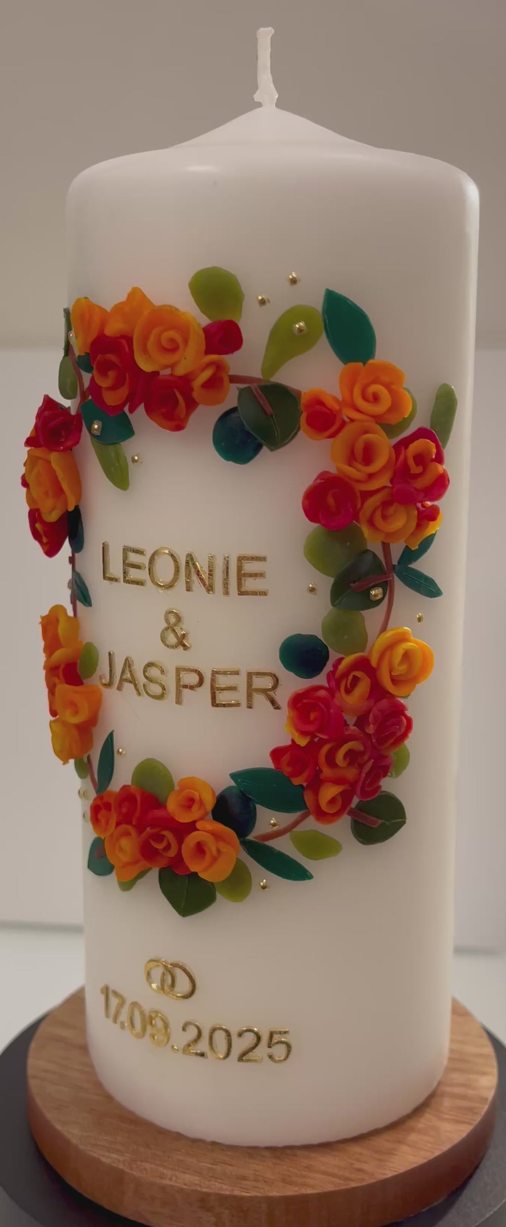 Weiße Hochzeitskerze mit handgefertigtem Wachsblumenkranz in Gelb, Orange und Rot, goldener Aufschrift „Leonie & Jasper“, goldenen Ringen und Hochzeitsdatum 17.09.2025, auf Holzsockel präsentiert.