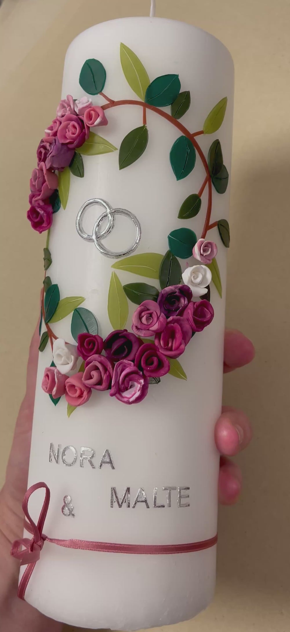 Weiße Hochzeitskerze mit handgefertigtem Wachsblumenkranz in Rosa-, Pink- und Lilatönen mit grünen Blättern, silbernen Ringen, silberner Aufschrift „Nora & Malte“ und rosa Schleifenband am unteren Rand.