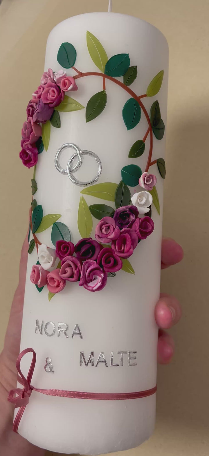 Weiße Hochzeitskerze mit handgefertigtem Wachsblumenkranz in Rosa-, Pink- und Lilatönen mit grünen Blättern, silbernen Ringen, silberner Aufschrift „Nora & Malte“ und rosa Schleifenband am unteren Rand.