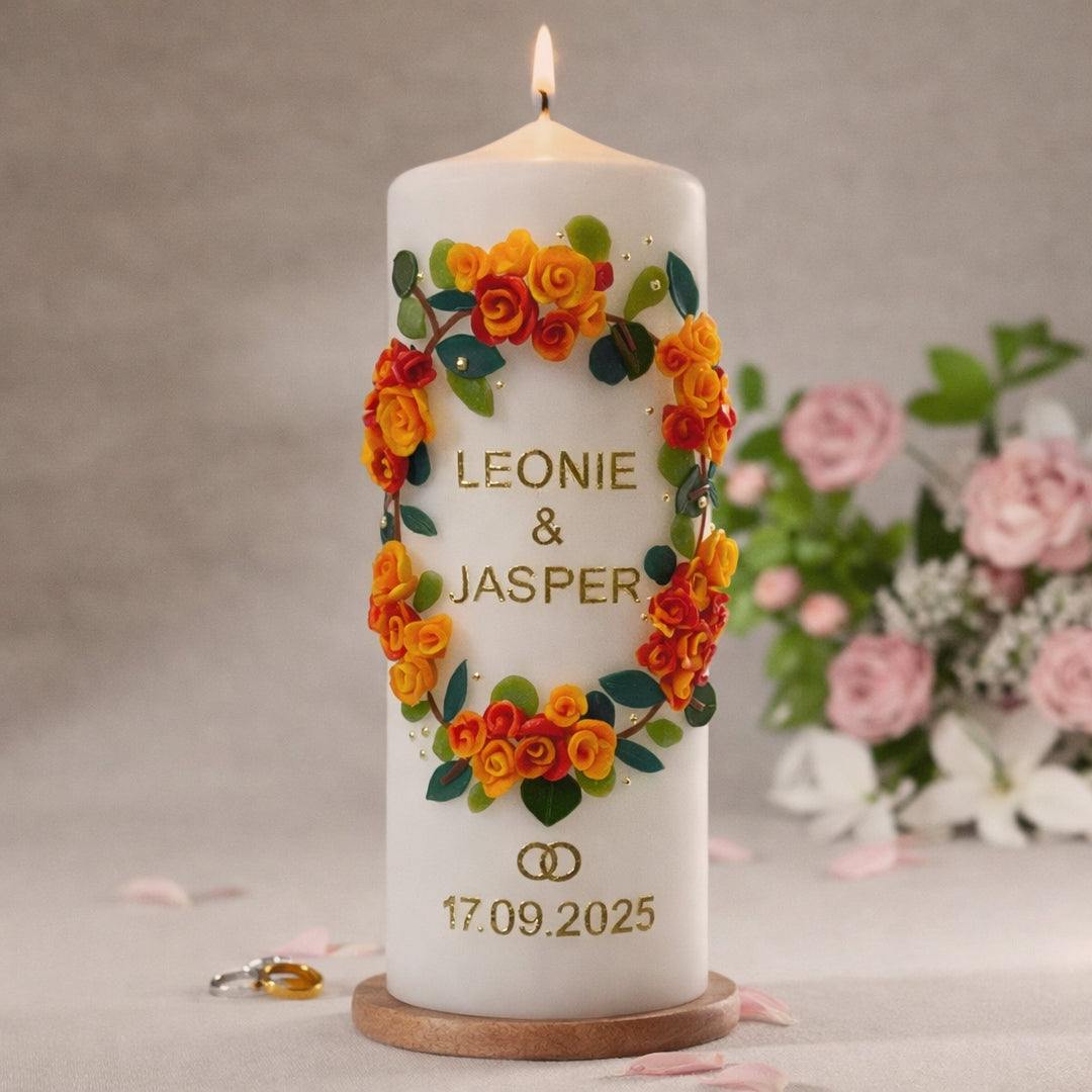 Weiße Hochzeitskerze mit handgefertigtem Wachsblumenkranz in Gelb, Orange und Rot, goldener Aufschrift „Leonie & Jasper“, goldenen Ringen und Hochzeitsdatum 17.09.2025, auf Holzsockel präsentiert.