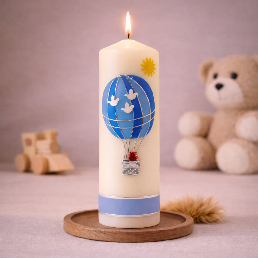 Elfenbeinfarbene Taufkerze mit blauem Heißluftballon und Teddy-Motiv, stehend auf Holzsockel vor neutralem Hintergrund mit Teddybär