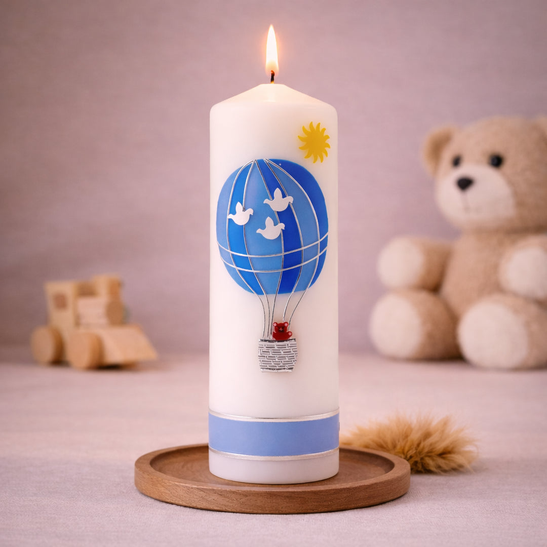 Weiße Taufkerze mit blauem Heißluftballon und kleinem Teddy im Korb, brennend auf Holzsockel vor neutralem Hintergrund mit Teddybär